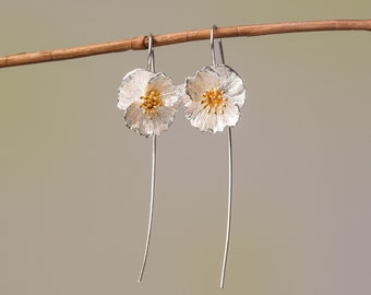 May include: Silver dangle earrings with a white flower design and gold accents. The flowers have a textured surface.