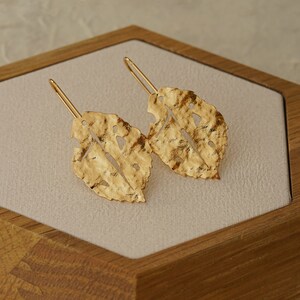 May include: A pair of gold leaf-shaped earrings with a textured finish. The earrings are hanging from a wooden display box.