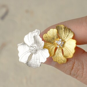 May include: A pair of flower-shaped earrings with a silver and gold finish. Each earring features a flower with five petals and a cluster of clear rhinestones in the center.