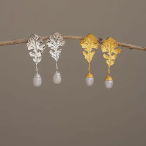 May include: Silver and gold-toned leaf earrings with pearl drops. The earrings are designed with detailed leaf shapes and feature a dangling pearl at the bottom. The earrings are displayed on a brown branch against a neutral background.