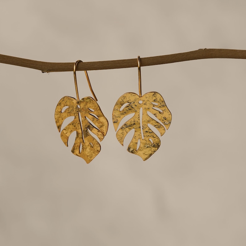 May include: Gold-tone earrings with a textured monstera leaf design. The earrings are hanging from a branch.