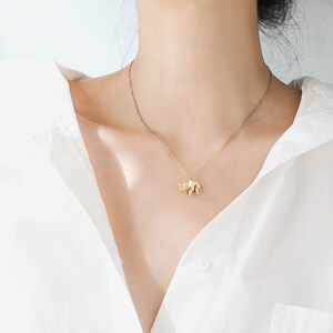 May include: A gold-colored necklace featuring a small elephant pendant. The elephant charm has detailed features and is suspended from a delicate chain. The necklace is worn against a white shirt.