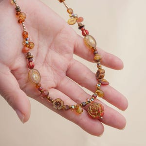 May include: A handcrafted necklace featuring a variety of amber, brown, and red beads. The necklace includes flower and leaf-shaped beads, along with a rose pendant. The beads are strung on a delicate gold-colored chain, creating a warm, earthy aesthetic.