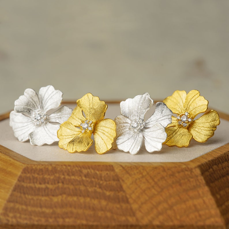 May include: A pair of flower-shaped earrings with a textured finish. One earring is gold and the other is silver. Each earring has a small clear crystal in the center.