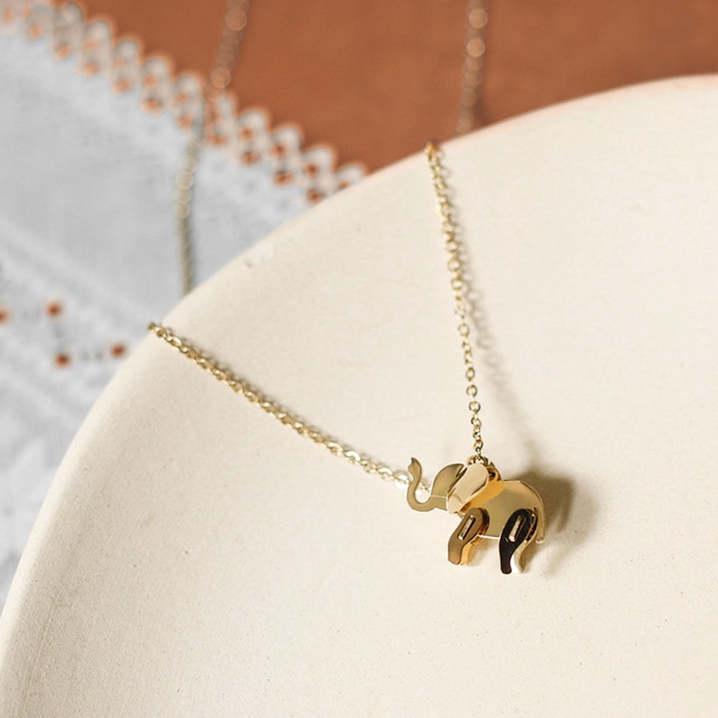 May include: A gold-colored elephant pendant necklace. The elephant charm is a geometric design with a trunk raised. The necklace chain is delicate and gold-toned. The necklace is displayed on a white, curved surface.