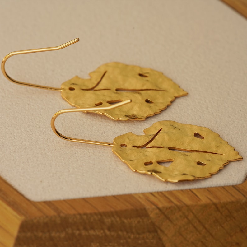 May include: A pair of gold leaf-shaped earrings with a hammered texture and cut-out details. The earrings have a simple hook closure.