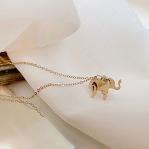 May include: A gold-colored necklace featuring a small, stylized elephant pendant. The elephant has detailed cutouts and is suspended from a delicate chain. The necklace is displayed on a white, draped fabric background.