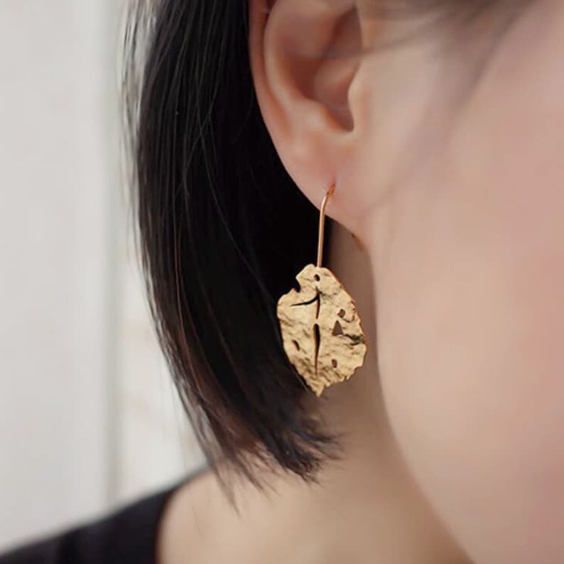 May include: Gold leaf earrings with a delicate, textured design. The earrings are shaped like a leaf with a hole in the center.