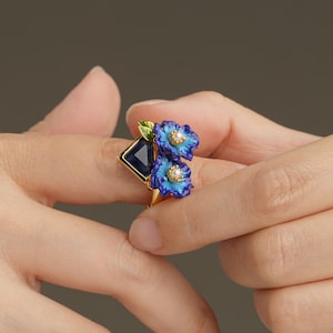 Pode incluir: Um anel de ouro com uma pedra preciosa azul escura e dois desenhos de flores azuis com detalhes brancos. O anel tem um design floral único com uma gema quadrada, sobre um fundo neutro.