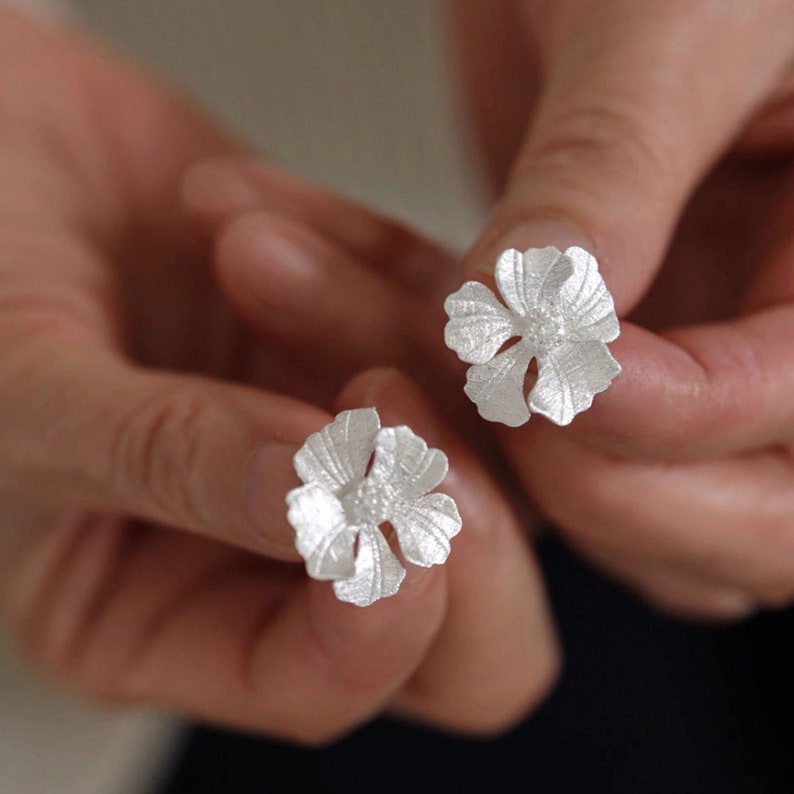 May include: A pair of silver flower-shaped earrings. The earrings are delicate and have a textured surface.