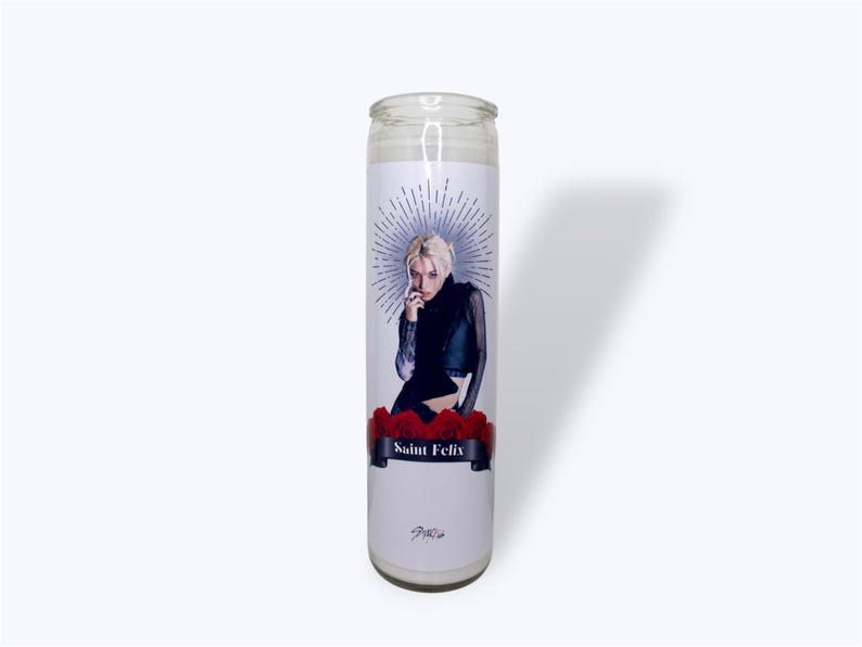 Peut inclure: Une bougie en verre blanc avec une image imprim&eacute;e d'une personne aux cheveux blonds portant une chemise noire et assise sur des roses rouges. Le texte "Saint Felix" est imprim&eacute; sur une banni&egrave;re noire sous l'image.