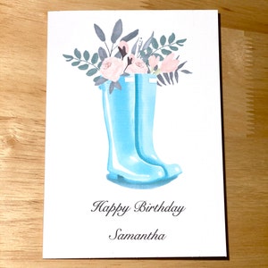 May include: A birthday card featuring a design of blue rain boots. The boots are filled with pink flowers and green leaves. The text "Happy Birthday Samantha" is printed on the card.