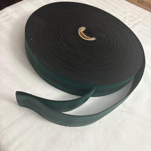 May include: A large, circular roll of dark forest green nylon webbing strap. The material features a tight, woven texture with a consistent, durable finish throughout the entire length of the spool, which is coiled tightly around a central cardboard core.