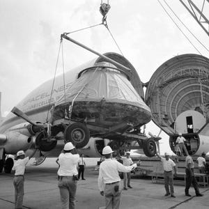May include: Black and white photograph of a large aircraft with its cargo bay open, revealing the interior structure. A covered object is being lifted into the plane by a crane. Several people in white shirts and hard hats are present.
