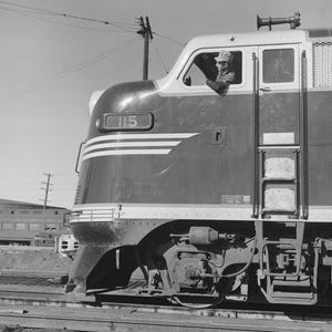 May include: A black and white side view of a diesel locomotive engine with the number 115 displayed on the front. The train features horizontal stripes on the nose, a ladder on the side, and a cab window where a person is visible looking out.