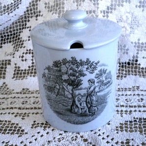 May include: A pale blue ceramic jar with a lid and a small handle. The jar has a black and white pastoral scene with figures and trees. The jar is on a white lace tablecloth.
