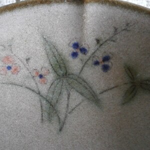 May include: A close-up of a ceramic bowl with a light brown glaze. The bowl is decorated with a hand-painted design of delicate flowers and leaves in shades of pink, blue, and green.