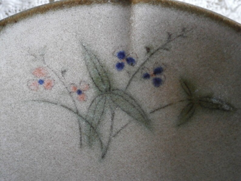 May include: A close-up of a ceramic bowl with a light brown glaze. The bowl is decorated with a hand-painted design of delicate flowers and leaves in shades of pink, blue, and green.