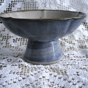 May include: A blue and white ceramic pedestal bowl with a scalloped rim. The bowl is sitting on a white lace tablecloth.