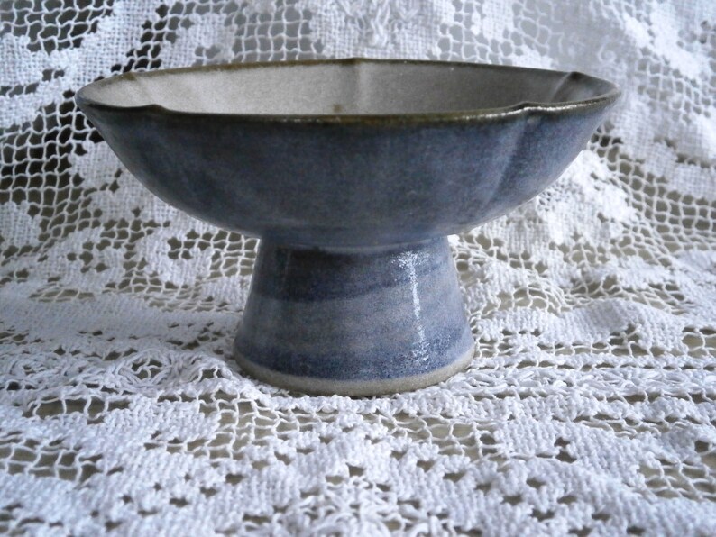 May include: A blue and white ceramic pedestal bowl with a scalloped rim. The bowl is sitting on a white lace tablecloth.