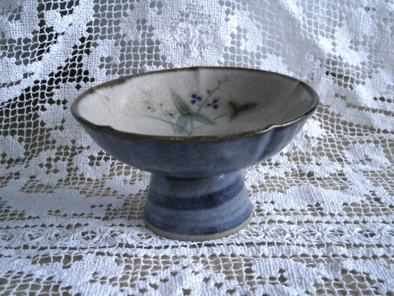 May include: A small, blue and white ceramic pedestal bowl with a floral design. The bowl has a scalloped edge and a raised base.