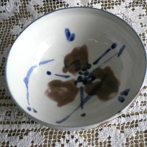 Peut inclure: Un bol en céramique blanche avec un motif abstrait bleu et brun. Le bol a une forme légèrement arrondie et une finition lisse et brillante.