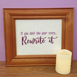 Puede incluir: Un punto de cruz enmarcado con el texto "Si no te gusta tu historia... Reescríbela" en letras moradas sobre un fondo blanco. El marco es de madera marrón y hay una vela blanca en primer plano.
