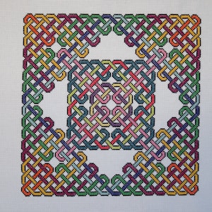 Op de afbeelding: Een kleurrijk Keltisch knoopontwerp geborduurd op witte stof. De knoop is gemaakt van meerdere garenstrengen in verschillende kleuren, waaronder rood, oranje, geel, groen, blauw, paars en roze.