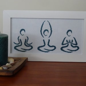 May include: A white framed cross-stitch with three blue figures in a yoga pose. The figures are stylized and simple.