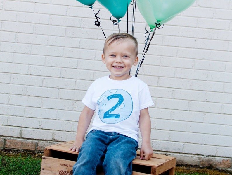 May include: A young boy wearing a white t-shirt with a blue number "2" on it, sitting on a wooden crate. He is smiling and looking at the camera. There are three green balloons behind him.