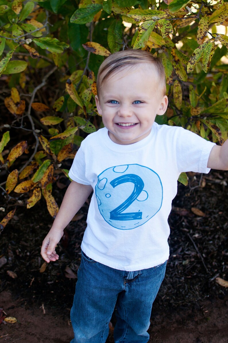 May include: A young boy wearing a white t-shirt with a blue moon and the number "2" on it. He is wearing blue jeans and smiling at the camera.