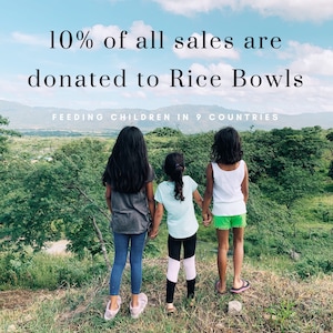 May include: Three girls stand on a grassy hilltop, looking out at a lush green landscape. The text "10% of all sales are donated to Rice Bowls FEEDING CHILDREN IN 9 COUNTRIES" is displayed at the top of the image.
