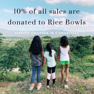 May include: Three girls stand on a grassy hilltop, looking out at a lush green landscape. The text "10% of all sales are donated to Rice Bowls FEEDING CHILDREN IN 9 COUNTRIES" is displayed at the top of the image.