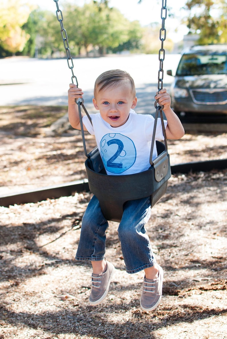 May include: A toddler wearing a white t-shirt with a blue moon graphic and the number "2" on it, sits on a swing set. The toddler is wearing blue jeans and brown shoes.