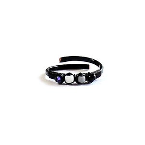 May include: A black metal ring with a curved design. The ring features a small purple stone, two white stones, and two black stones. The ring is set against a white background.