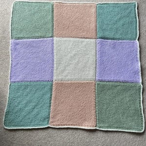 May include: A knitted patchwork blanket featuring nine squares in pastel shades of green, peach, lavender, and grey. The blanket has a white border and is displayed on a neutral grey surface. A cosy home decor item.