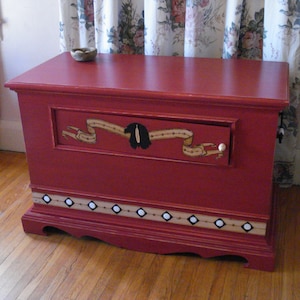 Op de afbeelding: Een rode houten opbergkist met een enkele lade en een decoratief geschilderd ontwerp. De lade heeft een witte knop en een geschilderd spandoek met een zwart vogelontwerp. De kist heeft een decoratieve rand met witte cirkels en zwarte vierkanten.