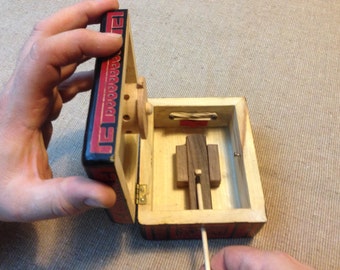 May include: A wooden box with a red and black design, open to reveal a small wooden mechanism with a wheel and a sliding piece. A wooden stick is inserted into the mechanism.