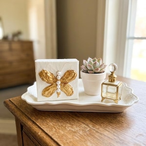 Puede incluir: Un lienzo rectangular blanco con una mariposa de concha de ostra dorada con cuerpo de perlas blancas, una pequeña suculenta en una maceta blanca y un frasco de perfume de vidrio transparente con marco dorado sobre una bandeja blanca.