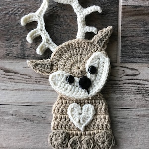 May include: A crocheted reindeer ornament with brown and white yarn. The reindeer has a white heart on its chest and black button eyes.
