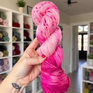 May include: A skein of yarn featuring a gradient of pink shades, transitioning from a pale, speckled light pink at the top to a vibrant, solid magenta at the bottom. The yarn has a twisted, multi-ply texture and is held in a vertical position.