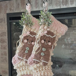 May include: Two handmade Christmas stockings, crocheted in cream, pink, and brown yarn. Each stocking features a gingerbread man applique and is adorned with faux greenery. The stockings hang from black metal hooks, perfect for holiday decor.