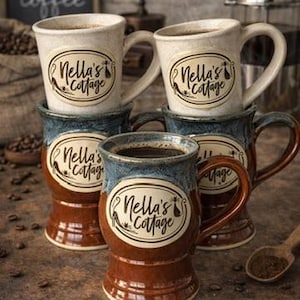 May include: A collection of ceramic coffee mugs with a two-tone design. The mugs are brown and blue with the text "Nella's Cottage" on the front. Coffee beans and a wooden spoon are also visible.