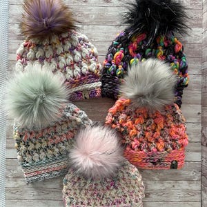 Peut inclure: Six bonnets en crochet colorés avec des pompons en fausse fourrure de différentes couleurs, dont le rose, le blanc, le gris, le noir et le violet.