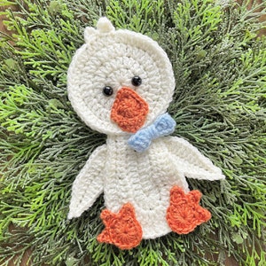 May include: White crocheted duck ornament with orange feet and a blue bow. The duck is sitting on a bed of green foliage.
