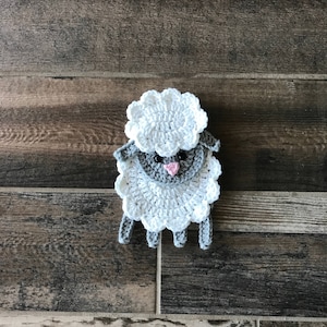 Op de afbeelding: Een gehaakt wit schaap met een grijs gezicht en een roze neus. Het schaap zit op een houten oppervlak.