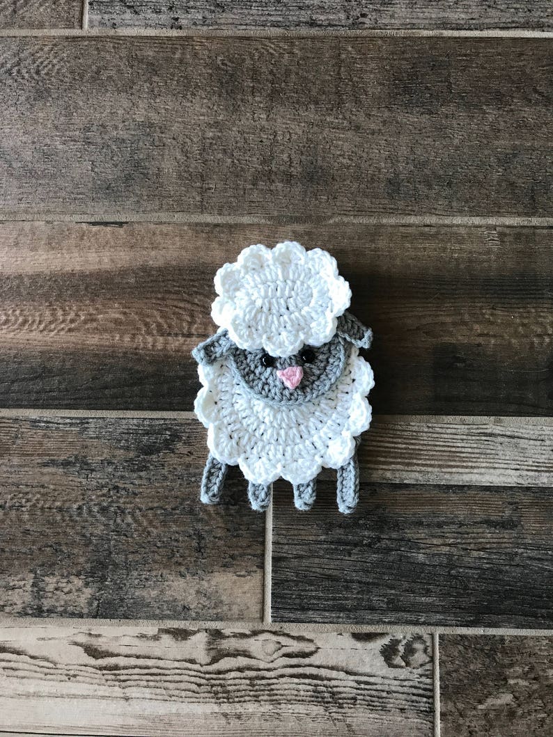 Op de afbeelding: Een gehaakt wit schaap met een grijs gezicht en een roze neus. Het schaap zit op een houten oppervlak.