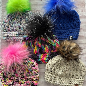 May include: Five handmade knit hats with faux fur pom-poms. The hats feature a variety of colors, including blue, pink, green, and neutral tones. Each hat has a unique knit pattern and a fluffy pom-pom on top. The hats are displayed on a light wood surface.