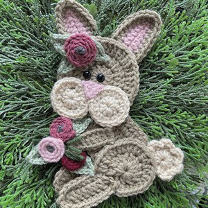 May include: A handmade crochet bunny with a floral crown and a bouquet of flowers. The bunny is primarily tan with pink accents in the ears and nose. The flowers are shades of pink and burgundy.