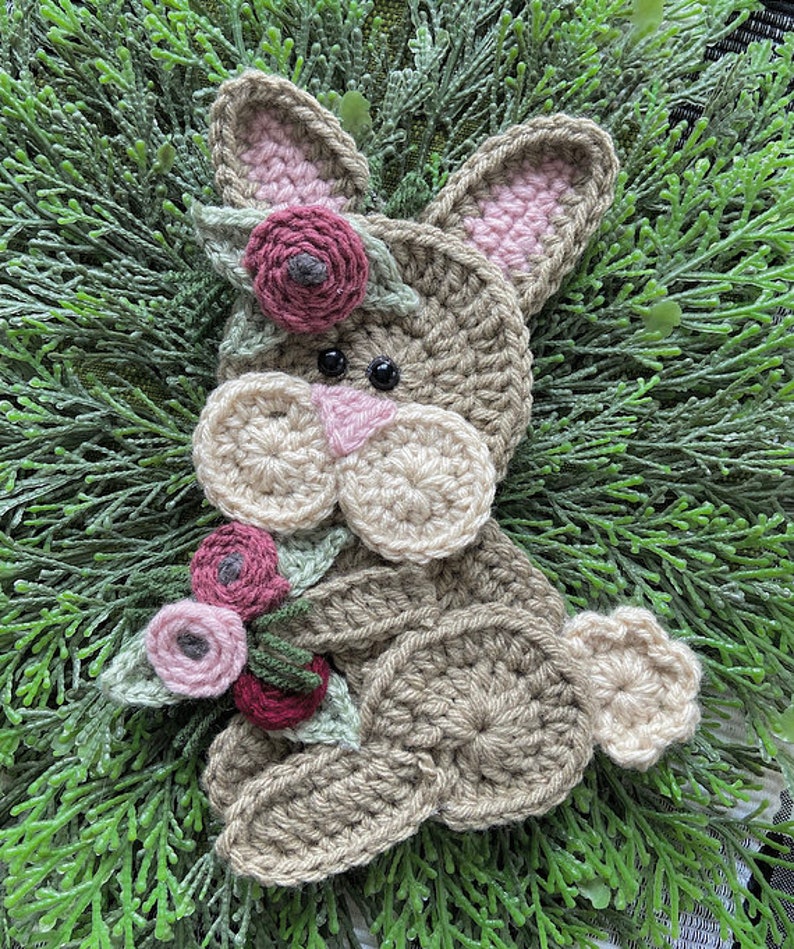 May include: A handmade crochet bunny with a floral crown and a bouquet of flowers. The bunny is primarily tan with pink accents in the ears and nose. The flowers are shades of pink and burgundy.